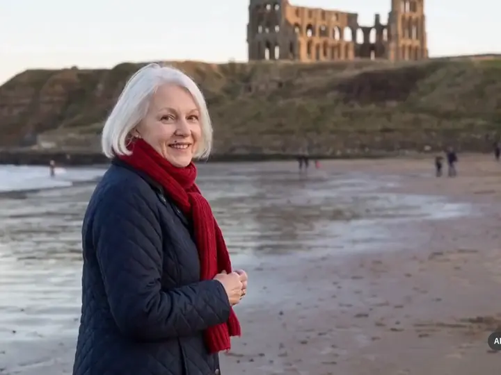 Eine weißhaarige Frau mit rotem Schal am Strand, hinter ihr die Ruine der Abtei auf der Klippe.